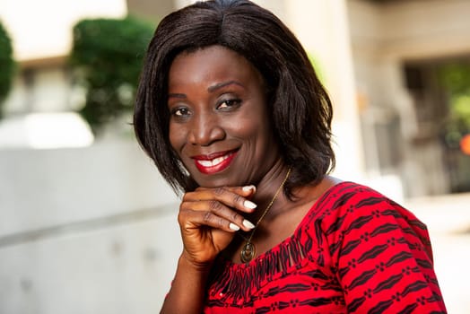outdoor portrait of a beautiful young African woman in a red loincloth outfit and with long hair, stands the mint with hand in the street looking at the camera,