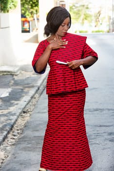 Stylish African woman in fashion, wearing a red loincloth and standing on a street while admiring and stroking her cloth worn on the shoulder,