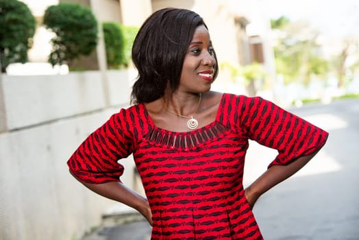 Stylish African woman in fashion, wearing a red loincloth and standing on a street, hands on hips looking at her side.