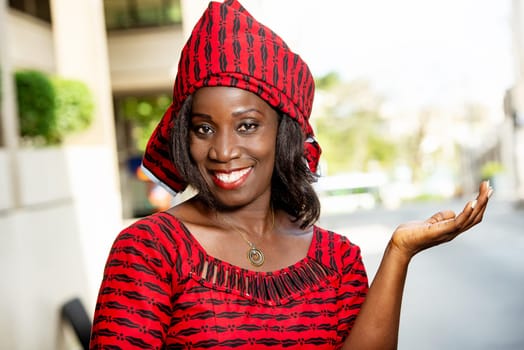mature african woman standing outdoors looking at camera smiling with outstretched hand.