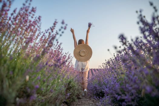 A woman is walking through a field of purple flowers with a straw hat on.