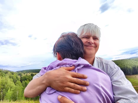 Two people share a warm hug against a backdrop of lush greenery and overcast skies in a peaceful outdoor setting.