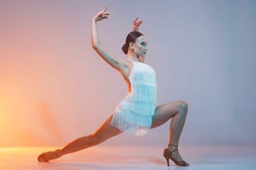 Elegant female Latin dancer in a white fringed dress performing a dynamic move with grace and confidence in artistic lighting.