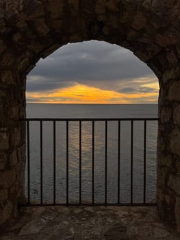 A rustic stone archway frames a view of calm blue waters and a golden sunset, with a black iron railing in the foreground. High quality photo. High quality photo