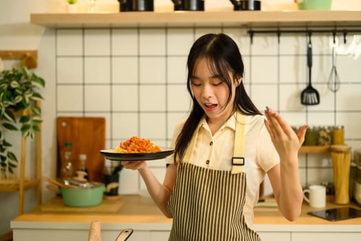 Young woman holding a freshly prepared plate of spaghetti with tomato sauce in her kitchen.