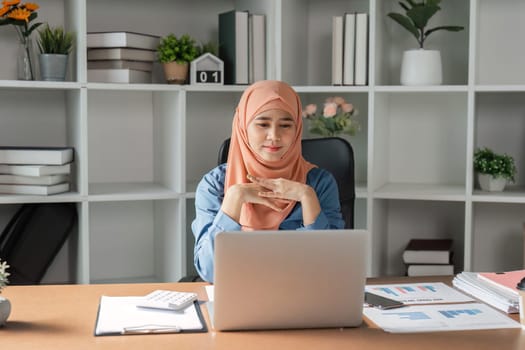 A Muslim woman wearing a hijab, attentively reviewing financial reports and data on her laptop in a contemporary office setting.