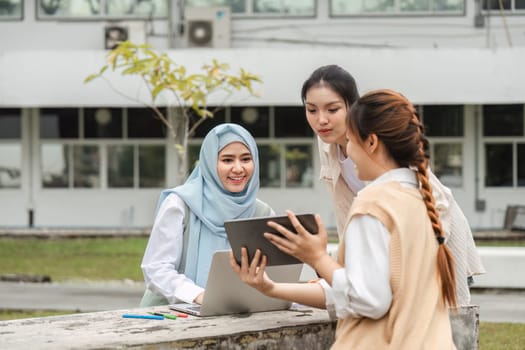 Three diverse female students engaged in a project discussion outdoors, utilizing a laptop and tablet in a collaborative learning environment.
