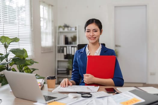 A happy businesswoman in a blue blazer proudly holds a red folder, showcasing her dedication to her financial career.