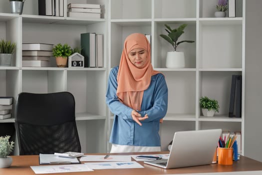 A diverse Muslim woman in a hijab showing discomfort in her wrist while seated at her office desk, highlighting the importance of ergonomic work practices.