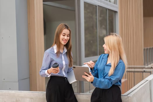 Two diverse women engaging in a discussion about work while holding a coffee and using a tablet in a modern office environment.