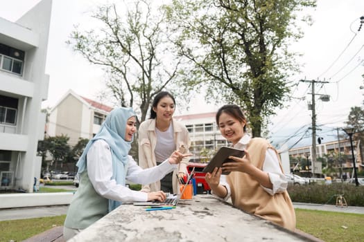 Three multicultural female students collaborating on a project outdoors, sharing ideas using a tablet and laptop in a vibrant campus environment.