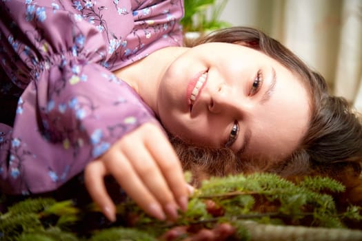 The young woman rests on leafy foliage, wearing a purple blouse, radiating joy and comfort in a cozy indoor setting.