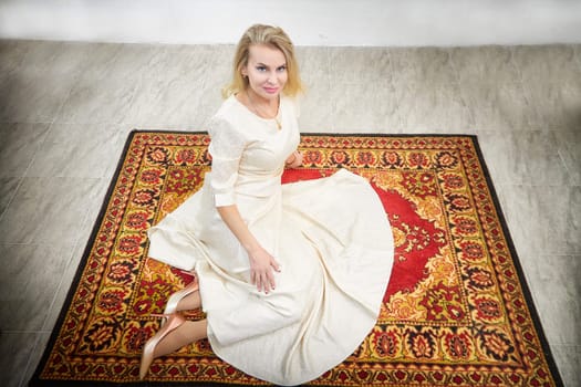 This middle-aged blonde blogger models in a stylish dress while posing on a decorative rug inside her home.