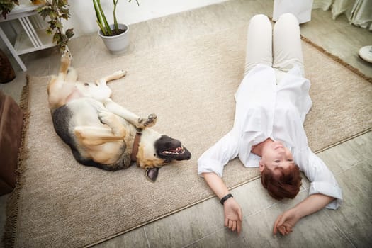 Middle-aged woman lies on the floor next to her happy German Shepherd in a warm, inviting room.