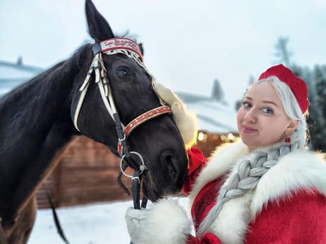 Russian girl in vibrant national dress interacts with a horse during the Maslenitsa holiday in a snowy landscape.