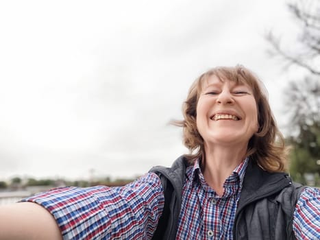 A middle-aged female traveler captures a cheerful selfie, enjoying nature during a trip in pleasant weather.