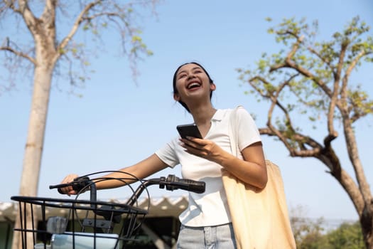 A joyful young woman smiles while holding her smartphone next to a bicycle, embodying a carefree outdoor lifestyle.