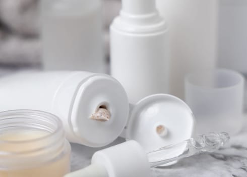 White opened cream tube with opened cap, pipette and jar on white marble table closeup. Beige scrub squeezing on table. Natural products for daily face care, spa procedures