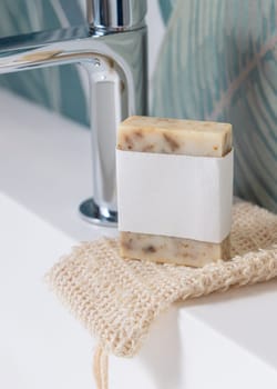 Beige handmade soap bar with blank label on sisal soap saver bag on washbasin close up, packaging mockup, copy space. Natural herbal unbranded product for daily hygiene