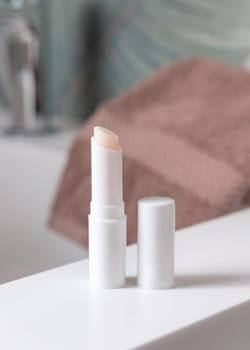 Opened white blank lip balm stick with a lid near brown towel on basin in green tropical bathroom close up, packaging mockup with copy space. Lifestile scene with ubranded natural lipcare product