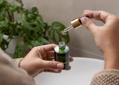 Female hands with natural manicure opening green cosmetic dropper bottle against green plant. Close up of woman in bathroom with drop of serum or essence falling from pipette