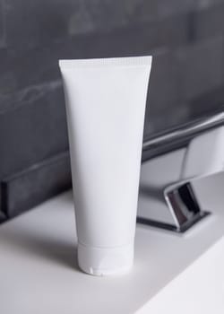 White cream squeeze tube near modern faucet on basin in dark grey masculine bathroom close up, packaging mockup. Natural unbranded cosmetics with copy space. Spa and wellness concept