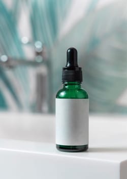 Green glass dropper bottle with pipette on washbasin with faucet in green tropical bathroom close up, label mockup. Natural unbranded cosmetics with copy space. Spa concept