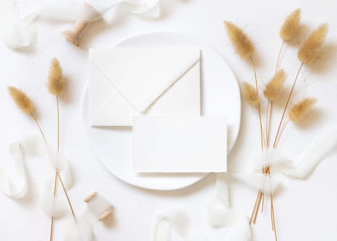 Blank card and envelope on white plate near curled silk ribbons and dried hare's tail grass topview, mockup. Pastel romantic flatlay. Bohemian or close to nature cards for party design or wedding stationery