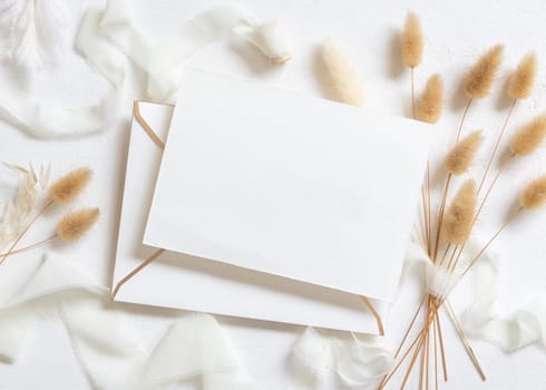 Blank card and envelope near curled silk ribbons and dried hare's tail grass topview on white, mockup. Pastel romantic flatlay. Bohemian or close to nature cards for party design or wedding stationery