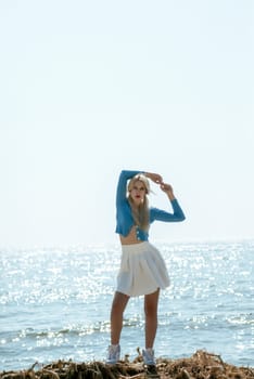 beautiful blonde girl stands on a sandy beach in sunny weather. girl in a short white skirt and a blue blouse. she stands against the backdrop of a calm sea. clear sea horizon.