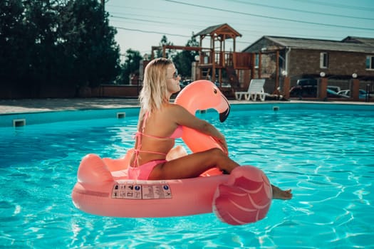 A woman is sitting on a pink flamingo float in a pool. The scene is bright and cheerful, with the woman enjoying her time in the water