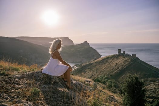 A woman is sitting on a hillside overlooking the ocean. She is wearing a white dress and has blonde hair. The scene is serene and peaceful, with the ocean in the background