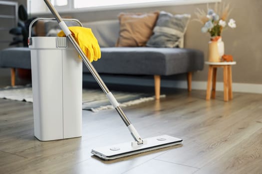 Cleaning supplies: mop, gloves, bucket with wringer sitting on wooden laminate floor in modern living room, wet equipment tools, housekeeping concept. Emphasizing home sanitation, cleanliness, hygiene