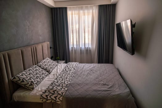 Modern bedroom interior: double bed with patterned bedding, wall-mounted TV, gray textured wall, and dark gray curtains flanking a window.