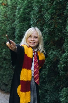 A young girl in a black robe and Gryffindor scarf holds a wand, smiling as she points it towards the camera.