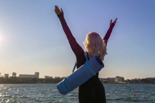 A woman with blonde hair stands with her arms raised, a yoga mat strapped to her back. She stands on a dock overlooking a body of water. The sky is blue and the sun is setting.