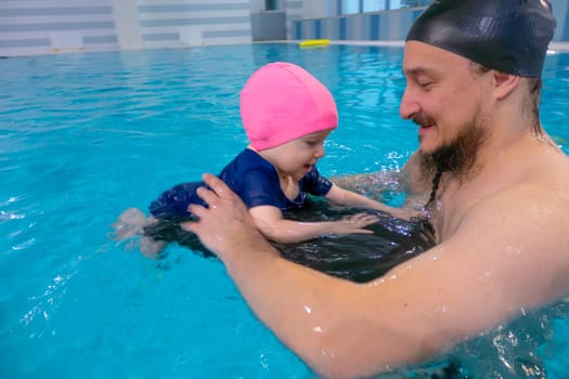 Happy middle-aged father swimming with cute adorable baby in swimming pool. Smiling dad and little child, girl having fun together. Active family spending leisure and time in spa hotel. Concept of kids sport, family summer vacation. Concept of healthy holiday and family activity