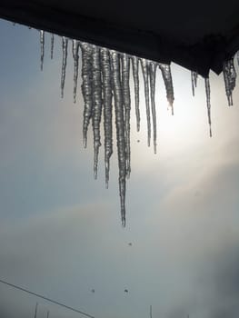 Melting icicles hanging from the roof of the balcony. Droplets of water falling from the sharp tips. The sun shining through the icicles.