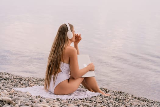 A woman is sitting on the beach with a laptop and headphones on. She is wearing a white bikini and she is enjoying her time by the water