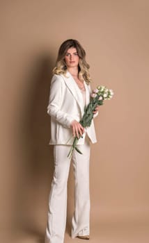 International Women's Day. Studio shot of a beautiful girl in a white pantsuit. The girl is standing on a beige background. She is holding flowers in her hands. She is looking at the camera.