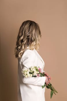 studio shot of a beautiful girl in a white pantsuit. the girl is standing on a beige background. she is holding flowers. profile portrait. her face is not visible, she is standing sideways