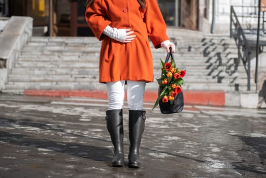 Closeup of woman in a orange coat and rubber boots walking down the street with a bouquet of tulips in spring city