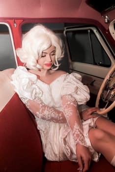 bright blonde in vintage lace white dress in stylish retro car. cosplay of Marilyn Monroe. girl has red lips and dark eyes, design inside the car, near the steering wheel. pin up style