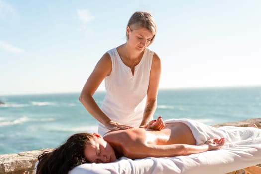 Therapist massaging woman's back on table at promenade by sea during sunny day. Professional masseuse is pampering female client during summertime
