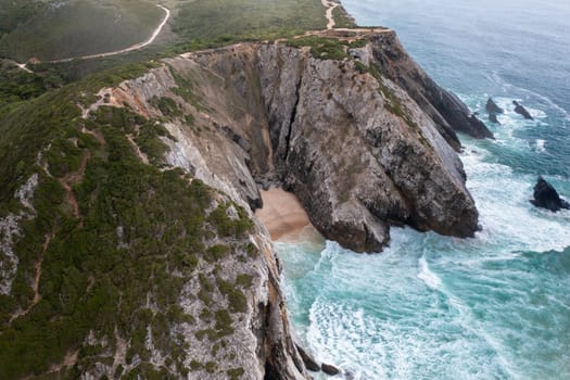 Drone view of massive rocky cliffs along seaside at scenic beach during daytime, surrounded by crashing waves, creating dramatic and picturesque coastal landscape