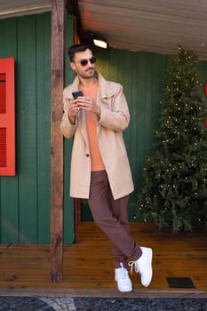Thoughtful man in beige trench coat holding smartphone outside green house. Full body of handsome young male is leaning on wooden pole during Christmas