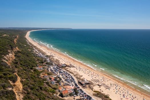 Aerial drone view showcases vibrant, crowded Fonte de Telha beach at Costa de Caparica, Portugal, with its golden sands, turquoise waters, and stunning seascape set against backdrop of blue sky
