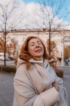 Red-haired woman, eyes closed, smiling serenely. Wrapped in a beige shawl outdoors, near bare trees and a grand building. Autumnal ambiance.
