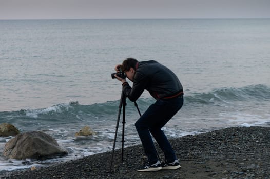A photographer bends over his camera, skillfully framing the serene ocean waves at dusk near rocky shores.