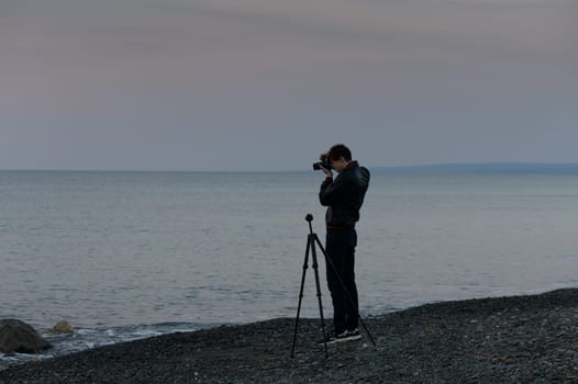 A photographer stands on a rocky beach at dusk, focused on capturing the serene waters under the soft evening light.